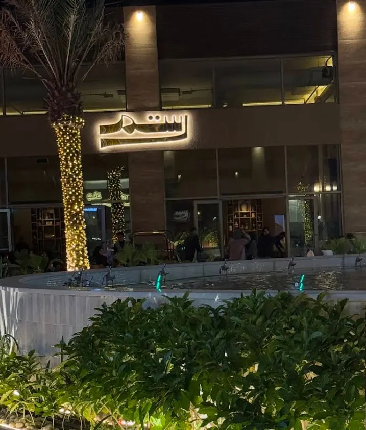 Rustom restaurant with its illuminated Arabic sign beside a lit palm and the plaza fountain